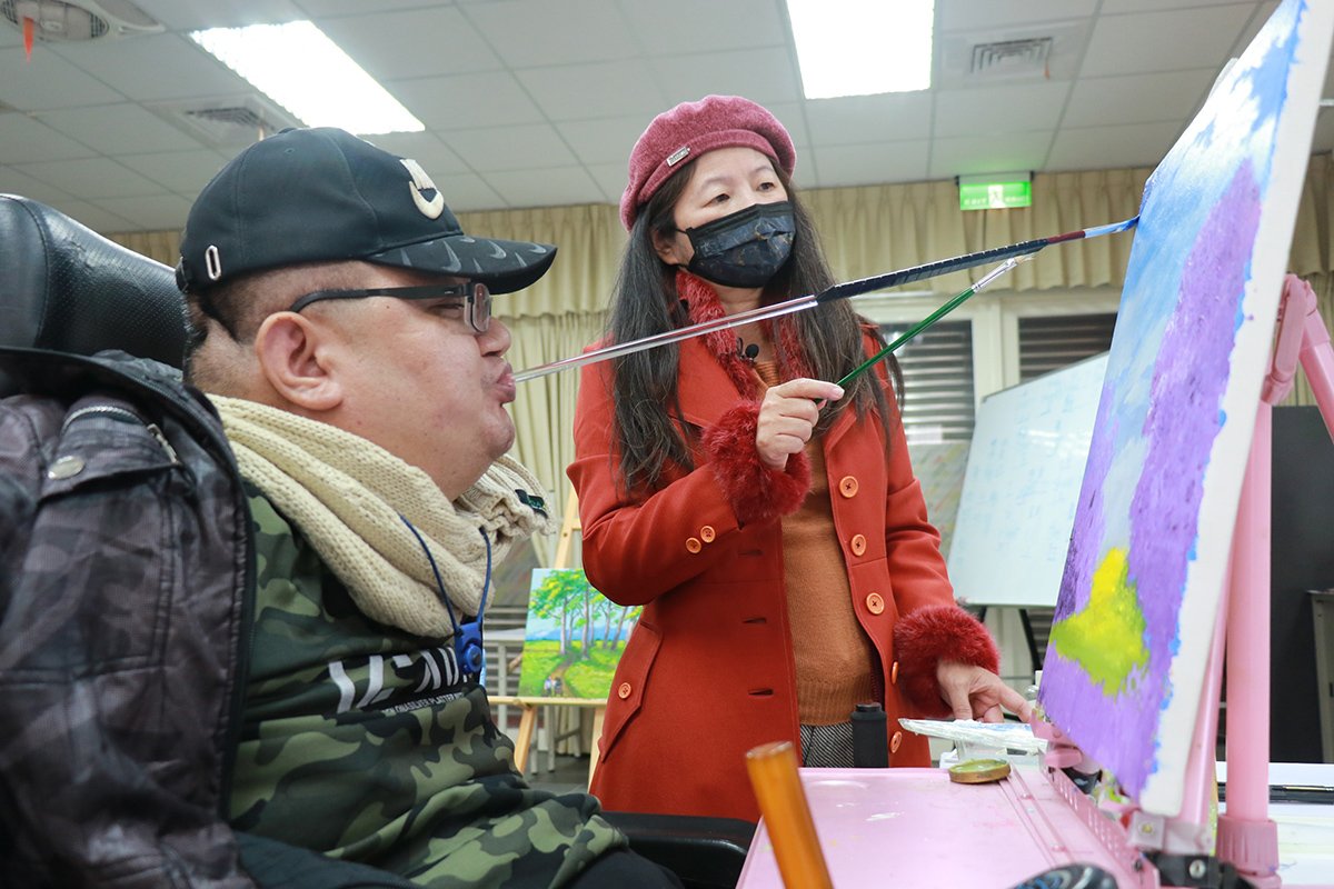 口足畫家江永旭畫卡感恩募愛 奉獻防疫與傷友 1 阮麗英老師(圖右)指導江永旭以口作畫。